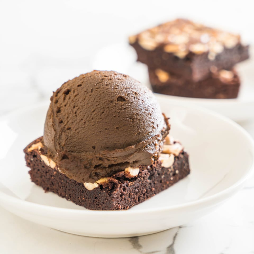 A scoop of chocolate ice cream sits atop a brownie with nuts on a white plate.