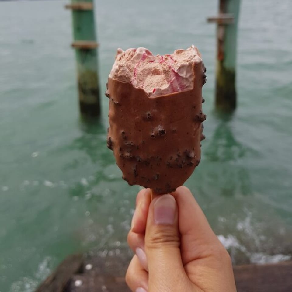 A hand holds a partially eaten chocolate-covered ice cream bar with a bite taken out, over water.